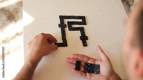 A close-up shot of a hand arranging black dominoes on a white surface, forming a creative pattern. The domino pieces are neatly laid out, and the blurred background adds focus to the action.