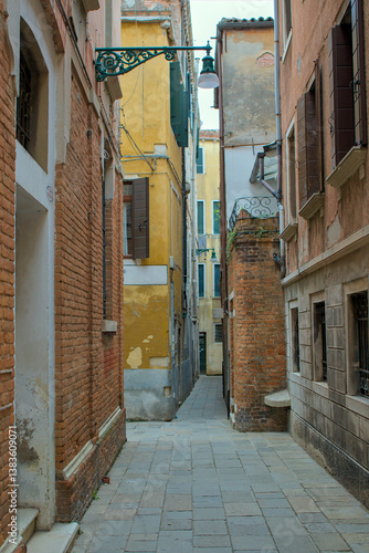 A narrow alley winds between old brick and stucco buildings in Venice, Italy. Weathered shutters and iron balconies give the scene a historic charm. A lone lamppost with ornate iron casts a soft glow.