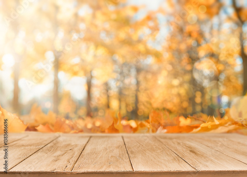 Autumn empty wooden table with falling yellow maple leaves on defocused autumn forest nature background at sunset. Thanksgiving, Halloween or autumn background with copy space.