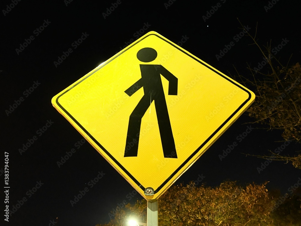 A yellow pedestrian crossing traffic sign with a black walking figure, illuminated against the night sky. A yellow pedestrian crossing traffic sign with a black walking figure, illuminated against the night sky.