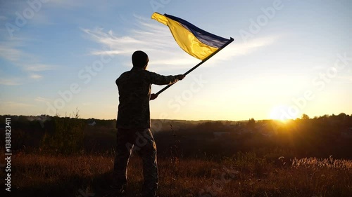 Ukrainian army man waving national banner against background of sunset. Young male soldier lifted flag at countryside. Victory against russian aggression. End of war between russia and Ukraine
