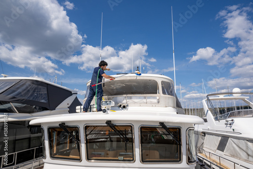 Boat maintenance worker providing cleaning service on a moored yacht. Maritime professional washing vessel deck with hose and brush at a scenic harbor.