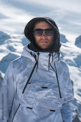 A man in a white and gray camouflage ski jacket with a hood and sunglasses stands in a snowy alpine environment with peaks and a partly cloudy sky.