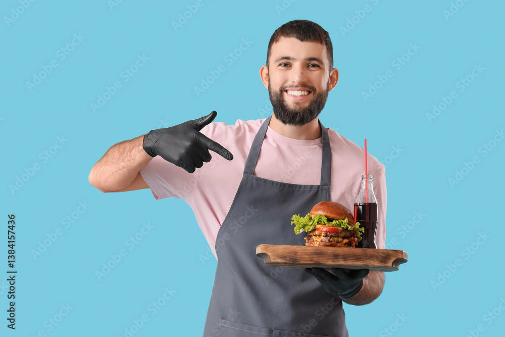 Young male chef pointing at wooden tray with tasty burger and cola on blue background Young male chef pointing at wooden tray with tasty burger and cola on blue background