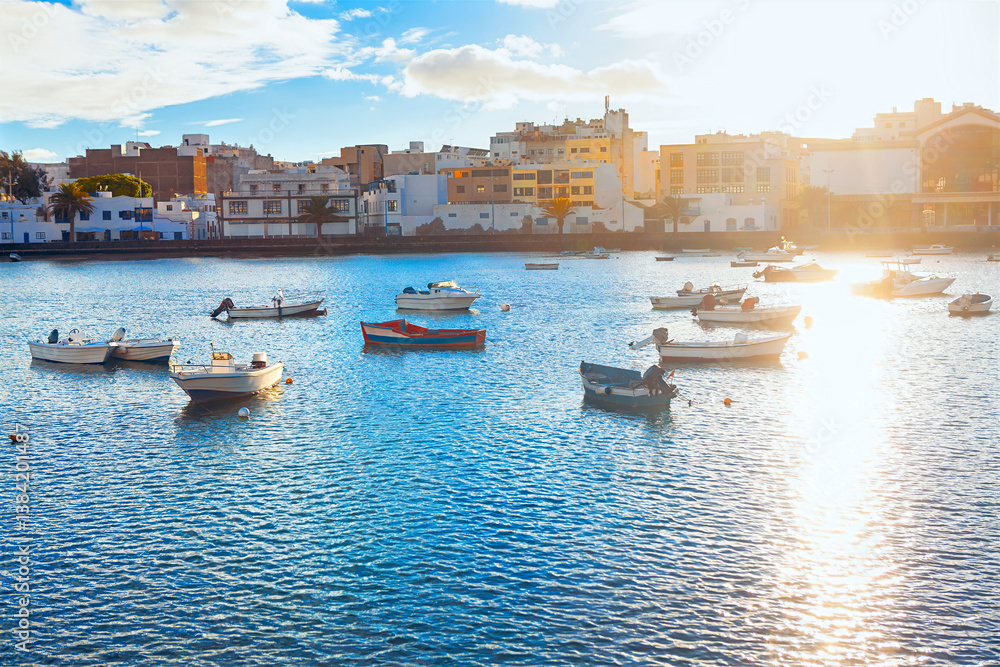 Sunlit coastal scene in Arrecife, Lanzarote, with small boats floating on calm waters. City buildings reflect the island's charm. Golden light adds warmth, creating a peaceful maritime atmosphere
