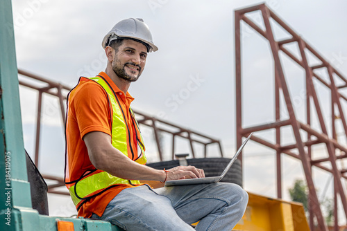 A male civil engineer in orange safety suit is using computer and application running a quality check of precast or cement floor products in a factory with crane background. Technology in a factory