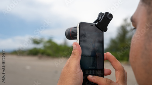 Billede på lærred A person capturing an outdoor landscape using a smartphone with an 18x telephoto lens attachment