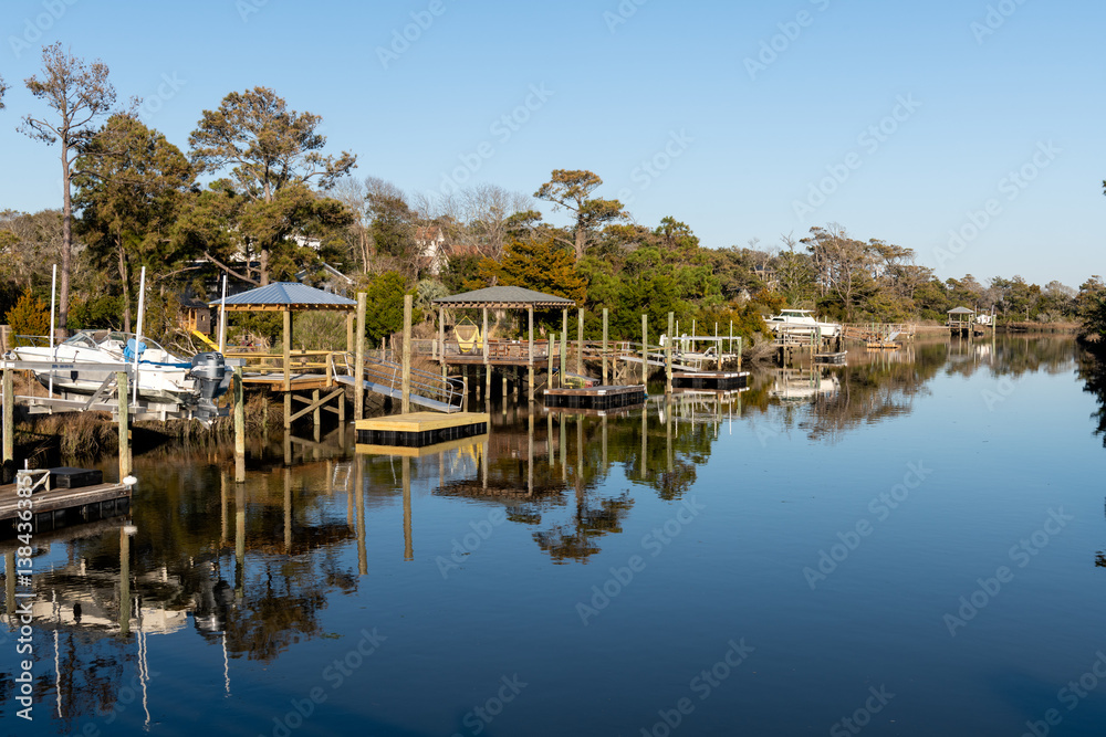 Fototapeta premium Docked boats reflect in the still waters of a peaceful canal lined with homes and trees in Oak Island, North Carolina.