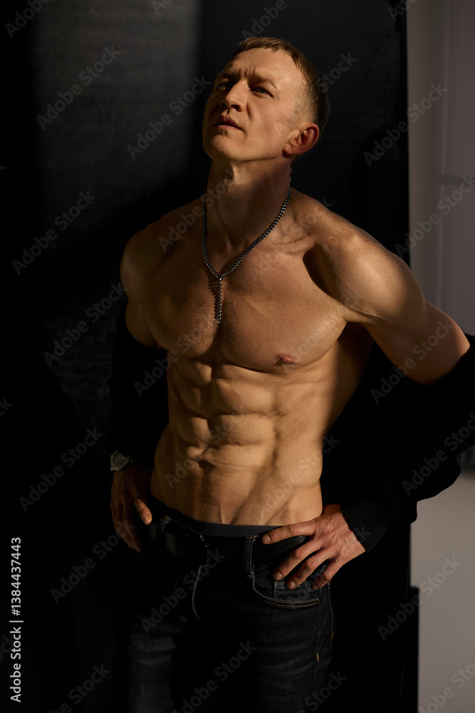 Fototapeta premium Slim muscular male model on black background. Shirtless fitness guy in black sweatpants posing in studio. Man with six pack abs in sunset light. Brutal photo