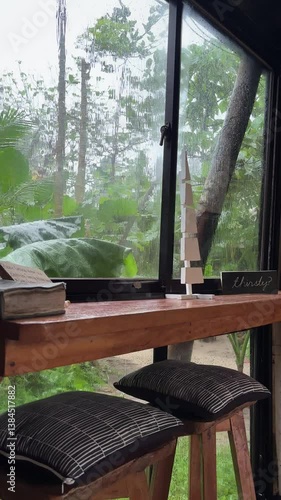 Warm wooden countertop with black/white cushions faces rainy window. Lush greenery visible through rain-streaked glass in peaceful cafe setting.
