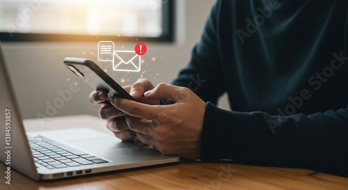 With a laptop on a wooden surface, a person uses a smartphone as an email notification icon with an exclamation mark appears above it.