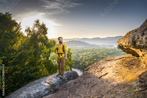 Bearded male hiker in a yellow shirt standing on a rocky cliff at sunset, overlooking a scenic valley with river, forest, and distant mountains.