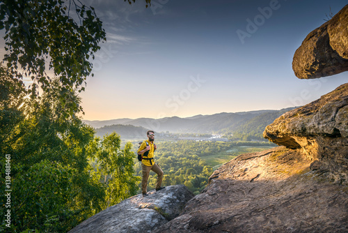 Bearded male hiker in a yellow shirt standing on a rocky cliff at sunset, overlooking a scenic valley with river, forest, and distant mountains.