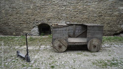 Electric scooter and medieval wooden cart against the backdrop of a stone fortress wall