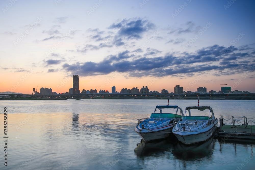 Fototapeta premium As the sun dips behind Taipei's skyline, Dadaocheng Wharf turns into a peaceful haven. Two boats rest quietly on the shimmering water, inviting serene moments under a dusky sky.
