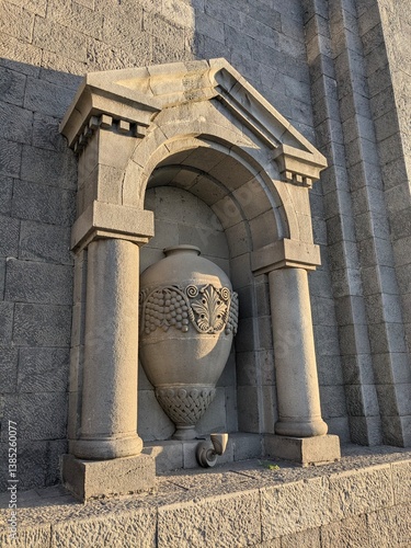 A large sculptural vase entwined with grapevine relief beneath a small classical portico, integrated into the historic architecture of the NOY Ararat Brandy Factory in Yerevan, Armenia
