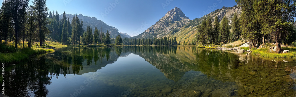 Fototapeta premium Panoramic photo of Lake. The lake is clear and calm, reflecting the surrounding mountains and trees in its surface. In front of it stands Mount Hood with snow-capped peaks against a blue sky backgroun