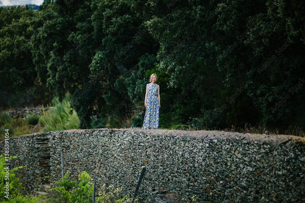 A woman in a summer dress stands on a stone wall in a vineyard in the Douro Valley, Portugal.