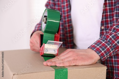Man using dispenser with adhesive tape to seal cardboard box indoors, closeup