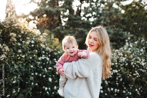 Smiling stylish woman holding baby girl 1 year old wearing knitted clothes over blooming bushes with flowers in meadow. Springtime. Motherhood. 