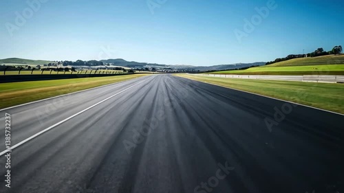 Smooth asphalt racetrack stretches into distance, surrounded by lush green hills and clear sky, evoking sense of speed and outdoor driving in natures serene landscape.