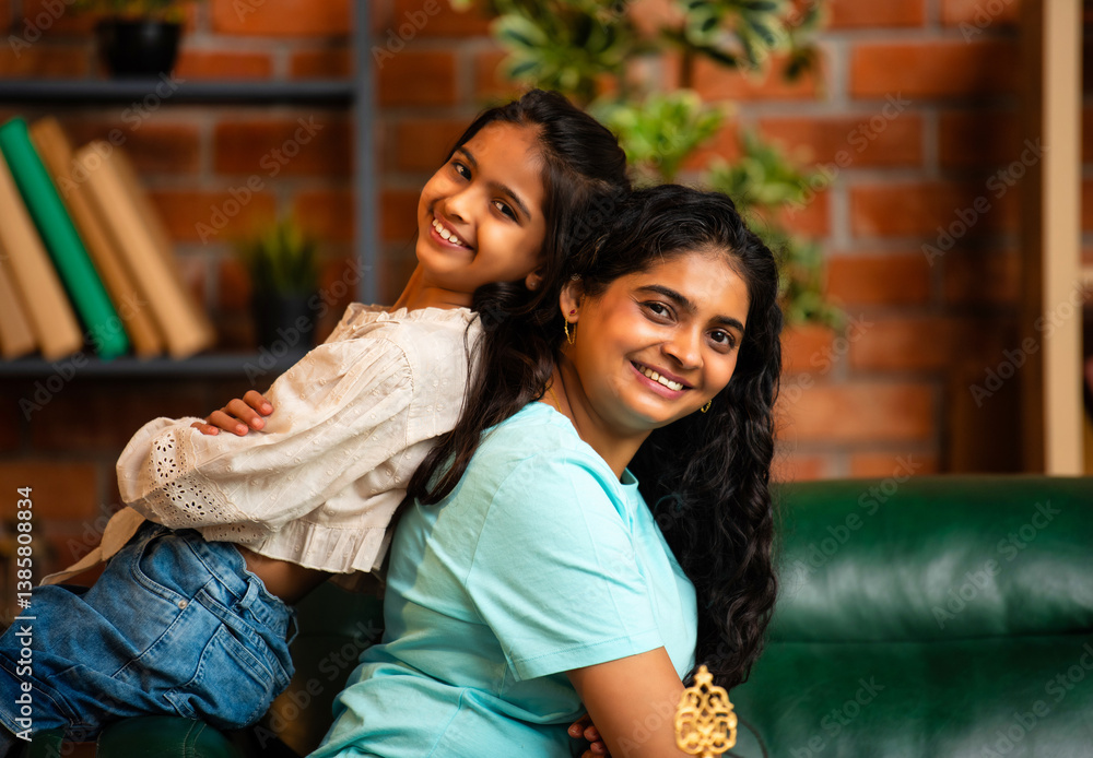 Fototapeta premium Playful Indian Asian mother and daughter sitting back to back, having fun on a comfy couch at home