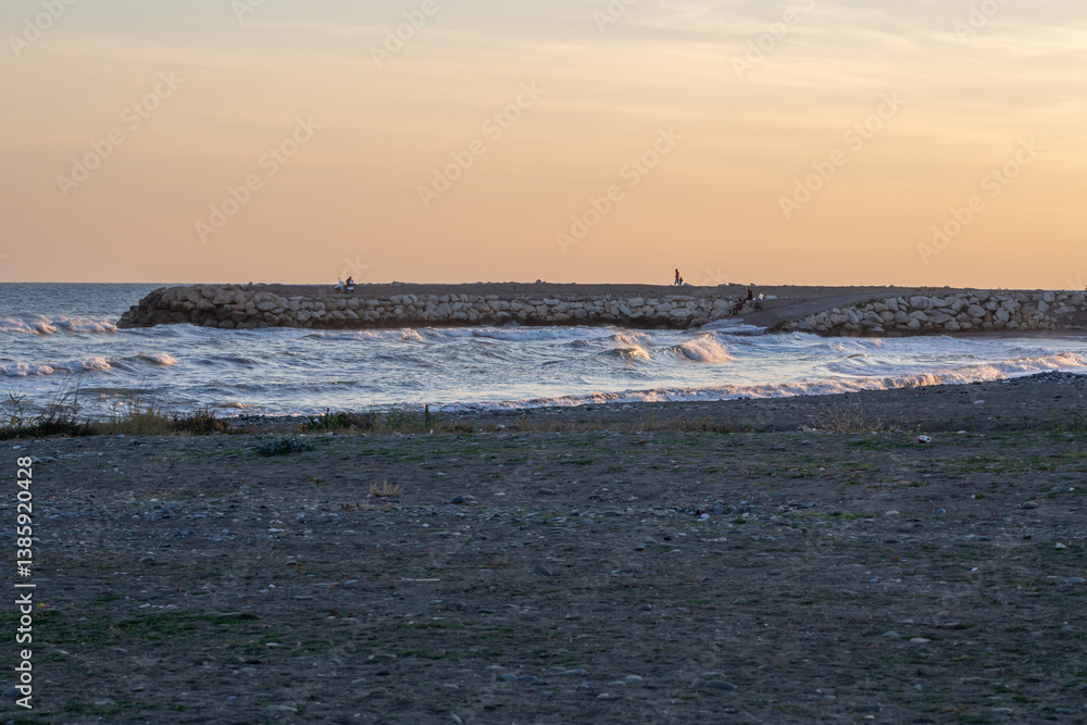 Waves gently crash onto the shore at sunset, reflecting soft golden light. A stone pier extends into the sea, where people sit in solitude, soaking in the peaceful ocean atmosphere. Waves gently crash onto the shore at sunset, reflecting soft golden light. A stone pier extends into the sea, where people sit in solitude, soaking in the peaceful ocean atmosphere.