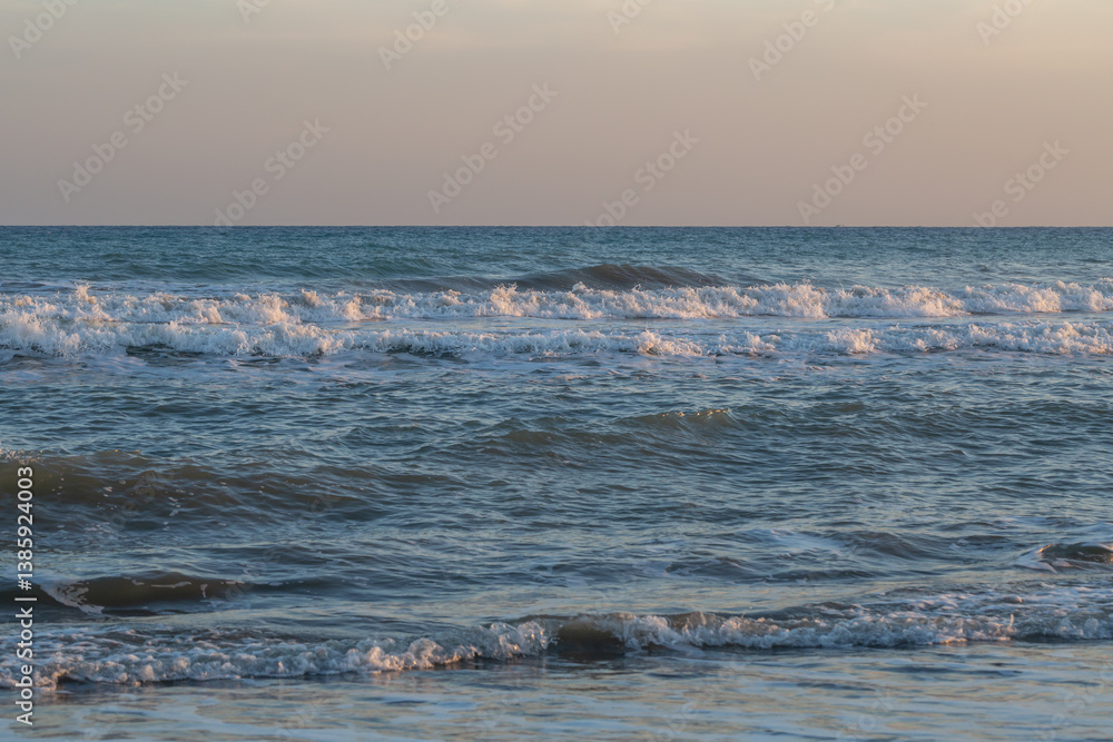 Obraz premium Crystal blue waves roll gently toward the shore under a pastel sky at sunset. The rhythmic movement of the sea creates a serene and calming atmosphere perfect for relaxation and reflection.
