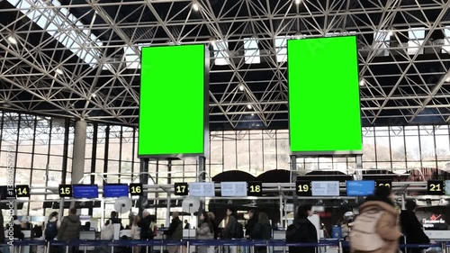 Blank green screen billboard in a bustling airport terminal, with blurred passengers moving through the space.
