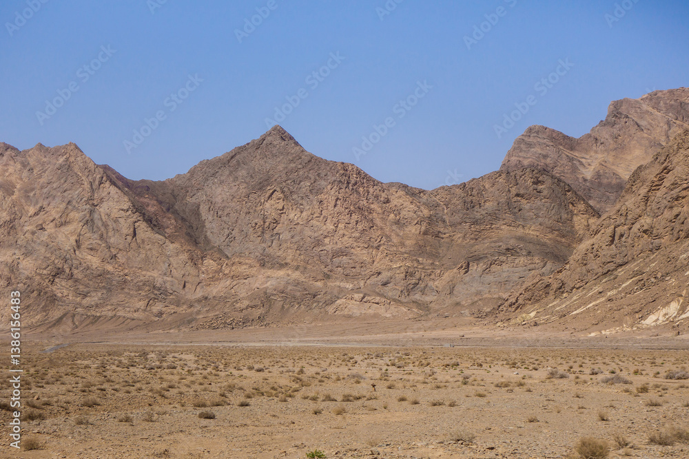 A high mountain range rises sharply from the plain. On the left is a wide strip of highway disappearing into the landscape. Shot in Yazd Province, Iran A high mountain range rises sharply from the plain. On the left is a wide strip of highway disappearing into the landscape. Shot in Yazd Province, Iran