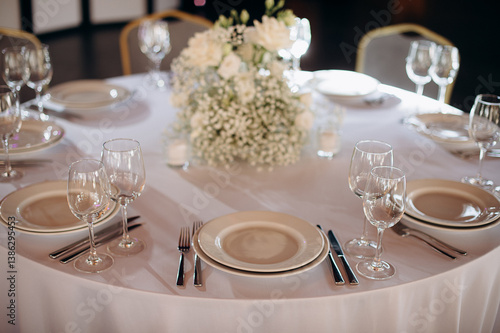 Elegant round wedding table setting with a white tablecloth, beige plates, simple white floral centerpiece, and tall wine glasses