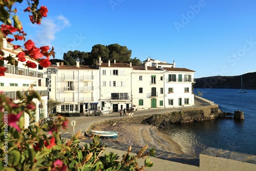 View of Cadaqués, a Mediterranean seaside town in springtime featuring small wooden boat on Es Pianc beach with people walking along promenade against backdrop of whitewashed buildings