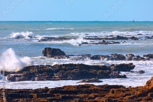Obraz na płótnie Powerful waves crash against jagged rocks, creating a dramatic coastal scene. vast expanse of ocean, dotted with dark rocks, stretches to the horizon w salonie