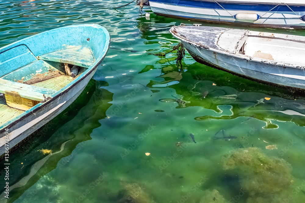 Obraz premium Two weathered boats float on the calm, greenish waters of Kotor's marina in Montenegro. Small fish swim beneath the boats, adding a peaceful ambiance to the coastal scene