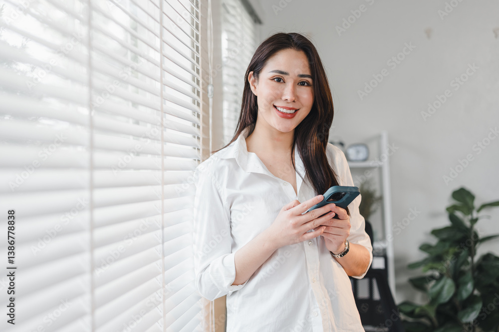 Obraz premium Cheerful woman in white shirt holding smartphone, standing by window with blinds in modern, bright interior.