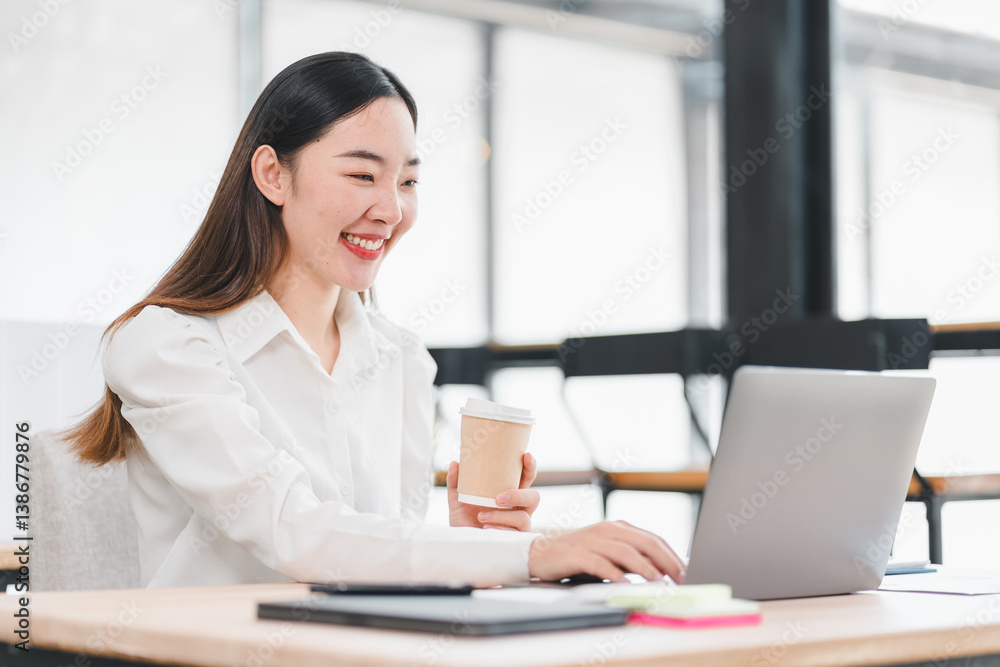 Cheerful woman in white shirt working on laptop while holding coffee cup in modern office setting. Cheerful woman in white shirt working on laptop while holding coffee cup in modern office setting.