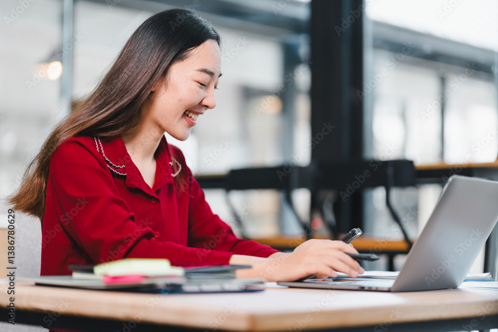 Woman in red shirt working on laptop at desk, smiling and focused in modern office setting. Woman in red shirt working on laptop at desk, smiling and focused in modern office setting.