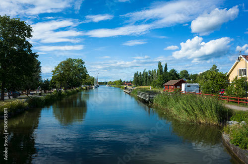 A scenic view of Göta Canal with calm waters reflecting a bright blue sky and white clouds. Lush greenery, traditional Swedish houses, and a peaceful summer atmosphere. Töreboda, Sweden