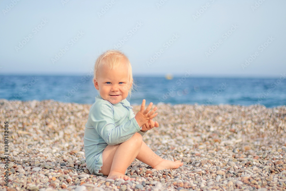 A joyful and cheerful toddler joyfully sits on a pebbly beach, smiling brightly with the vast ocean in the background