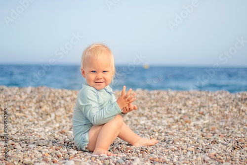 Obraz na płótnie A joyful and cheerful toddler joyfully sits on a pebbly beach, smiling brightly with the vast ocean in the background w salonie