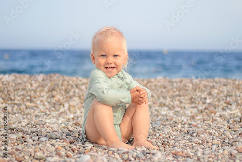 Obraz na płótnie A joyful and cheerful toddler joyfully sits on a pebbly beach, smiling brightly with the vast ocean in the background w salonie