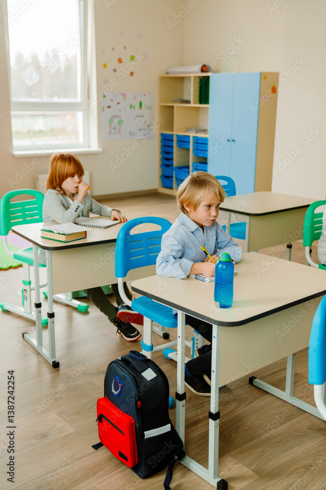 Obraz premium Two elementary school White students sit at desks and work on their studies in a classroom. The room contains colorful furniture, shelves, decorations, backpacks, and school supplies.