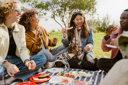 During a picnic in a sunny park, a multiethnic group of casually dressed young friends in their 20s sits laughing and eating fruit and bread on a blanket, with a dachshund sitting with an Asian woman.