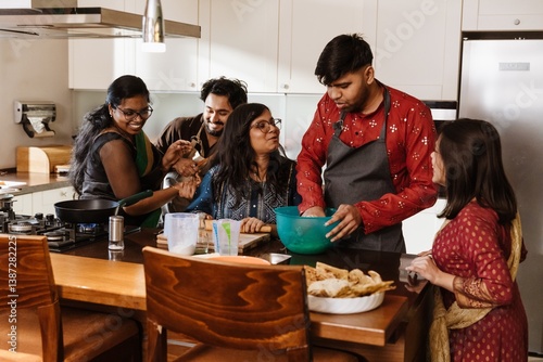 A happy Indian family gathered at a table in the kitchen with white cabinets and cooking dinner together, a man in a red shirt kneading the dough with two women, a woman in eyeglasses heats the pan