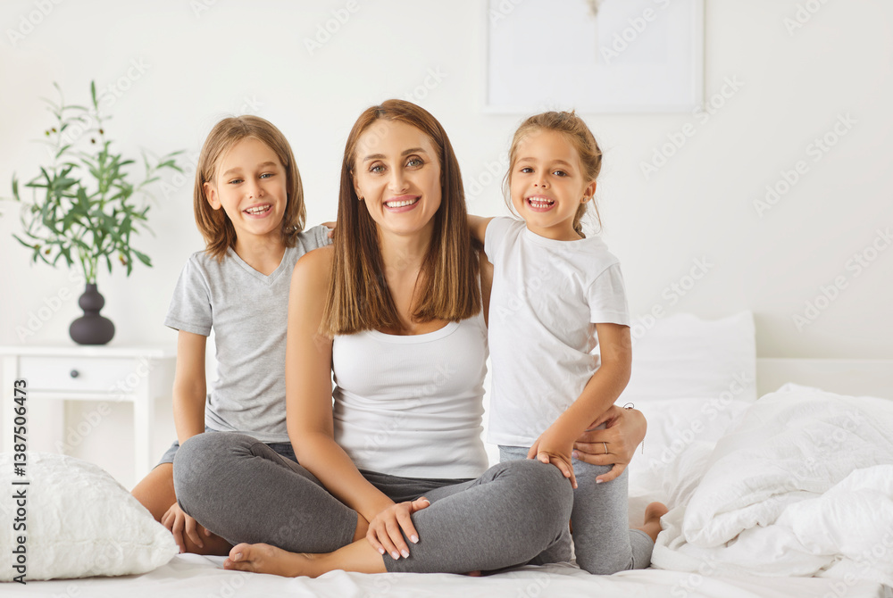 Portrait of happy smiling young mother with two children sitting on bed in bedroom at home in the morning and looking at camera enjoying spending time together. Family and mothers day concept Portrait of happy smiling young mother with two children sitting on bed in bedroom at home in the morning and looking at camera enjoying spending time together. Family and mothers day concept
