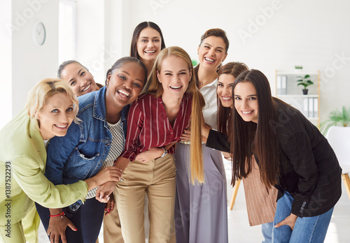 Group portrait of happy diverse women standing together in office and hugging each other. Happy female colleagues or friends laughing and embracing, showing unity and teamwork in positive atmosphere.