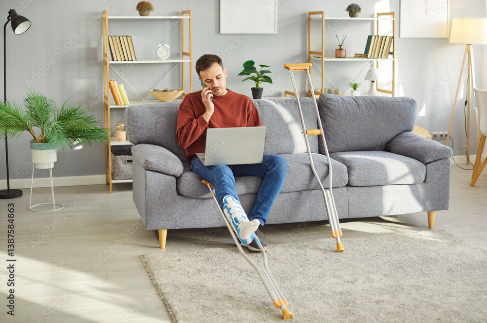 Injured young man with bandaged leg sitting on sofa, working on laptop and talking on mobile phone with crutches lying nearby. Male person recovering from foot trauma during remote work from home.