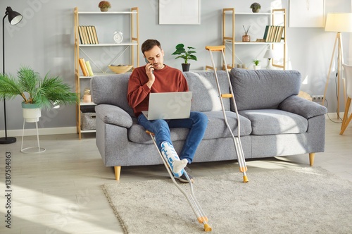 Obraz na płótnie Injured young man with bandaged leg sitting on sofa, working on laptop and talking on mobile phone with crutches lying nearby. Male person recovering from foot trauma during remote work from home. w salonie