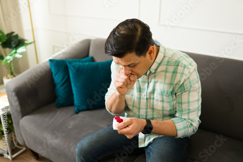 Bild auf Leinwand Feverish latino male coughing, wiping bloody sputum while seated on living room