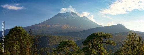 Popocatépetl volcano eruption smoke at the daytime among other green hills and mountains, with cloudy blue sky